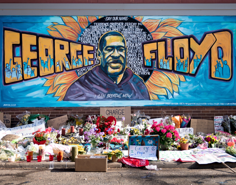 Photograph of the George Floyd Memorial in Minneapolis, Minn. A large blue and yellow mural on a brick wall featuring an image of George Floyd as well as his name in large letters and the words "I can breathe now." A collection of flowers, candles, and other memorial offerings are arranged in front of the mural. Source: Lorie Shaull via Creative Commons.