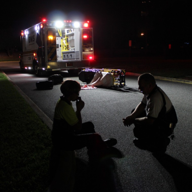 Image description: Two emergency medical technicians crouch in front of an open ambulance on a roadway at night time. There is an empty gurney visible in the background. Source: Creative Commons