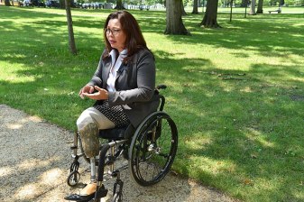 Senator Tammy Duckworth sits in her wheelchair on a sidewalk in front of a green lawn. She is wearing black glasses, a grey blazer, a white dress shirt, and a black and white polka dotted skirt. Source: Wikimedia Commons