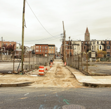 A street in North Baltimore, Md. with a badly damaged sidewalk. A series of orange construction barrels mark an uneven segment of alleyway pavement between two curb cuts that is filled with dried mud and would likely be impassible to many people using assistive devices. Parking lots and brick row homes stand in the background.  Image: Max Pixel, CC