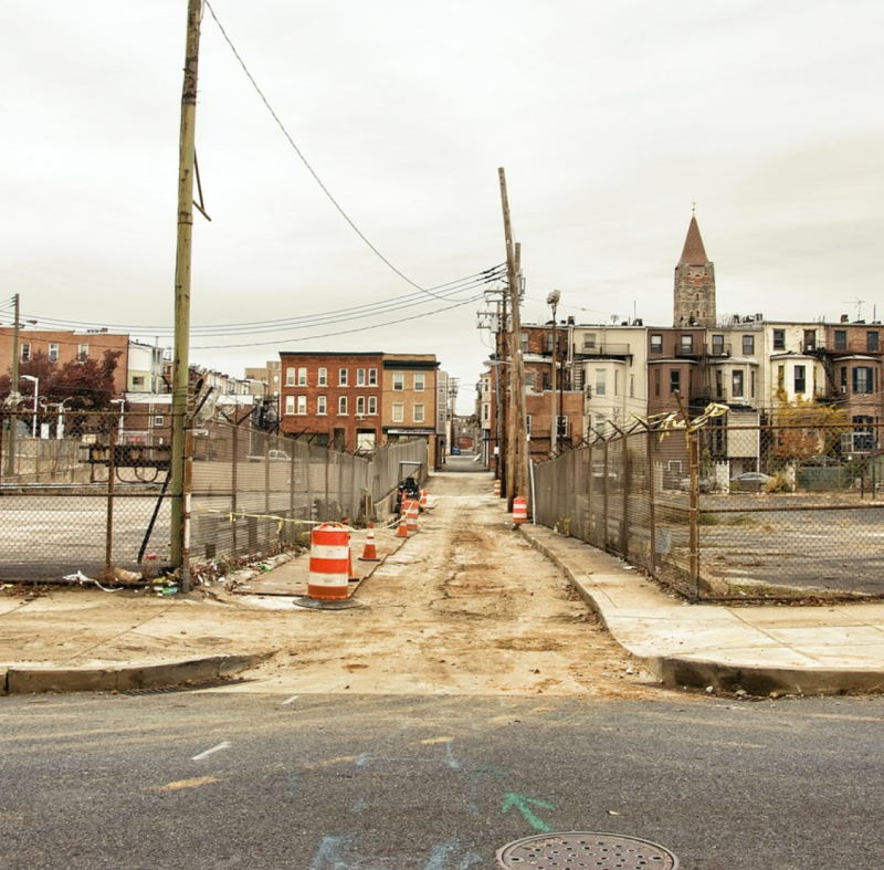 A street in North Baltimore, Md. with a badly damaged sidewalk. A series of orange construction barrels mark an uneven segment of alleyway pavement between two curb cuts that is filled with dried mud and would likely be impassible to many people using assistive devices. Parking lots and brick row homes stand in the background. Image: Max Pixel, CC