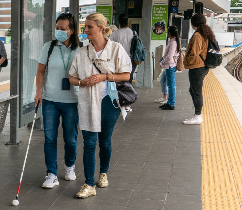 Image description: A person with a white cane and long dark hair walks on an outdoor train platform, accompanied by a friend. Photo: John Robert McPhereson, CC